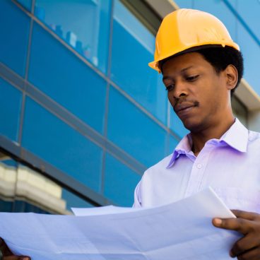 Portrait of professional architect in helmet looking at blue prints outside modern building. Engineer and architect concept.